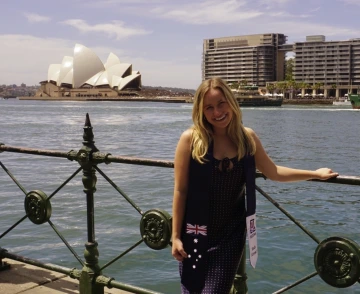 A smiling young woman stands by a waterfront railing with the iconic Sydney Opera House and a modern high-rise building in the background. She wears a dark dress with white polka dots and a navy stole featuring the Australian flag and the University of Arizona Study Abroad logo. The sunny day highlights the blue sky and calm water, suggesting a joyful moment during a study abroad experience in Sydney, Australia.