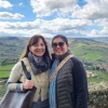Two women stand smiling in front of a scenic overlook with a sweeping view of a green countryside dotted with fields, trees, and small buildings under a partly cloudy sky. Both women wear layered winter clothing—one with a cream-colored vest and gray scarf, and the other in a black jacket with a patterned shoulder strap. They appear to be enjoying a peaceful moment in a rural, likely European, setting, possibly Italy. A stone wall and bench are visible in the foreground.