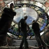 Three technicians in hard hats and work gear stand inside the vast, high-tech interior of the ATLAS detector at CERN’s Large Hadron Collider. The circular structure behind them is a massive, intricate assembly of wires, electronics, and machinery, with a large metallic disc at the center labeled “ATLAS CERN LHC.” The image conveys the scale and complexity of particle physics research and engineering. The lighting highlights the equipment, while the technicians appear slightly blurred, emphasizing motion and